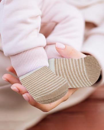 Beige knitted baby booties with ribbed texture and ankle cuffs shown against soft fabric background. White socks visible on infant's feet.