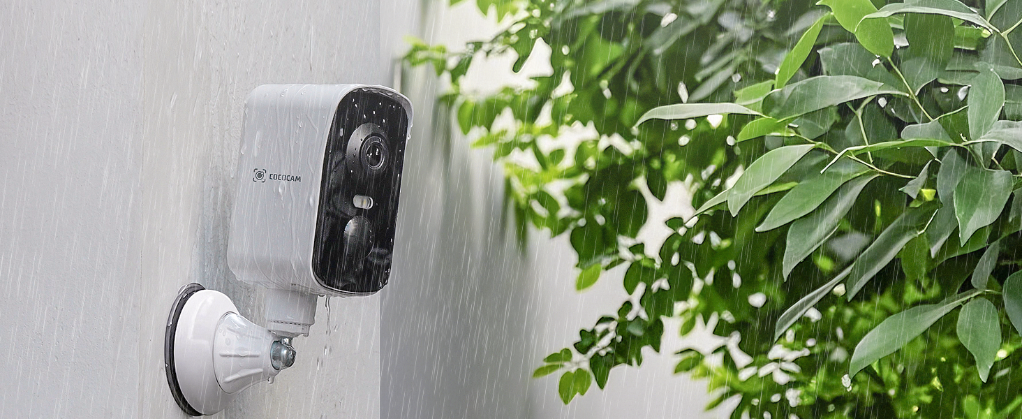 Outdoor security camera mounted on white wall. Black lens in white casing. Surrounded by green leaves, suggesting exterior placement for home monitoring.