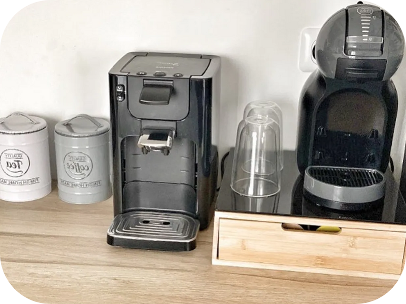 Stazione da caffè con due macchine da caffè, una grigia e una nera, insieme a tre contenitori bianchi su una superficie di legno