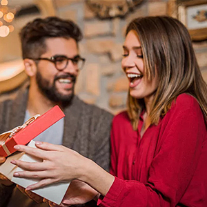 Woman in red blouse holding a wrapped gift box, smiling with excitement. Man with glasses beside her, also smiling.