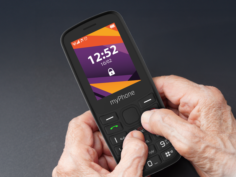 Mobile phone displaying time '12:52' on screen with orange-purple gradient background. Hands holding basic feature phone with keypad.