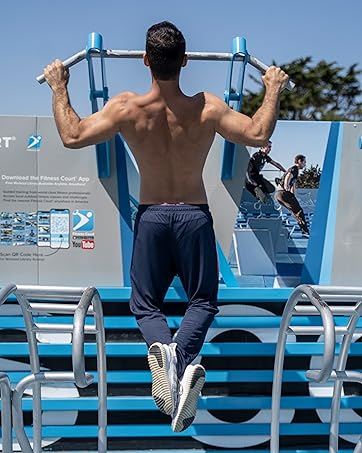 Shirtless man performing pull-up on outdoor exercise equipment. Blue sky and other exercise structures visible in background.
