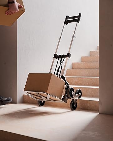 Zusammenklappbare Sackkarre im Einsatz auf Treppen. Schwarzer Metallrahmen mit zwei Rädern. Die Beine der Person sind sichtbar, wenn eine Pappschachtel eine Treppe hinauf bewegt wird.