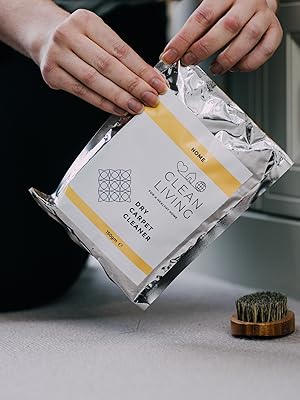 Hands opening a foil package or sachet against a gray surface, with a small brush visible in the frame.