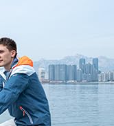 Una chaqueta de invierno azul y naranja que se muestra sobre un fondo de paisaje urbano frente al mar con edificios altos visibles al otro lado del agua