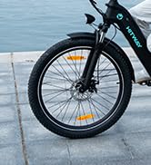 Primer plano de la rueda delantera de una bicicleta con freno de disco. Neumático negro con llanta amarilla visible contra un pavimento de color claro