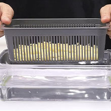 Hands holding a gray plastic container with vertical slots, filled with what appears to be pasta or noodles. The container sits above a clear surface.
