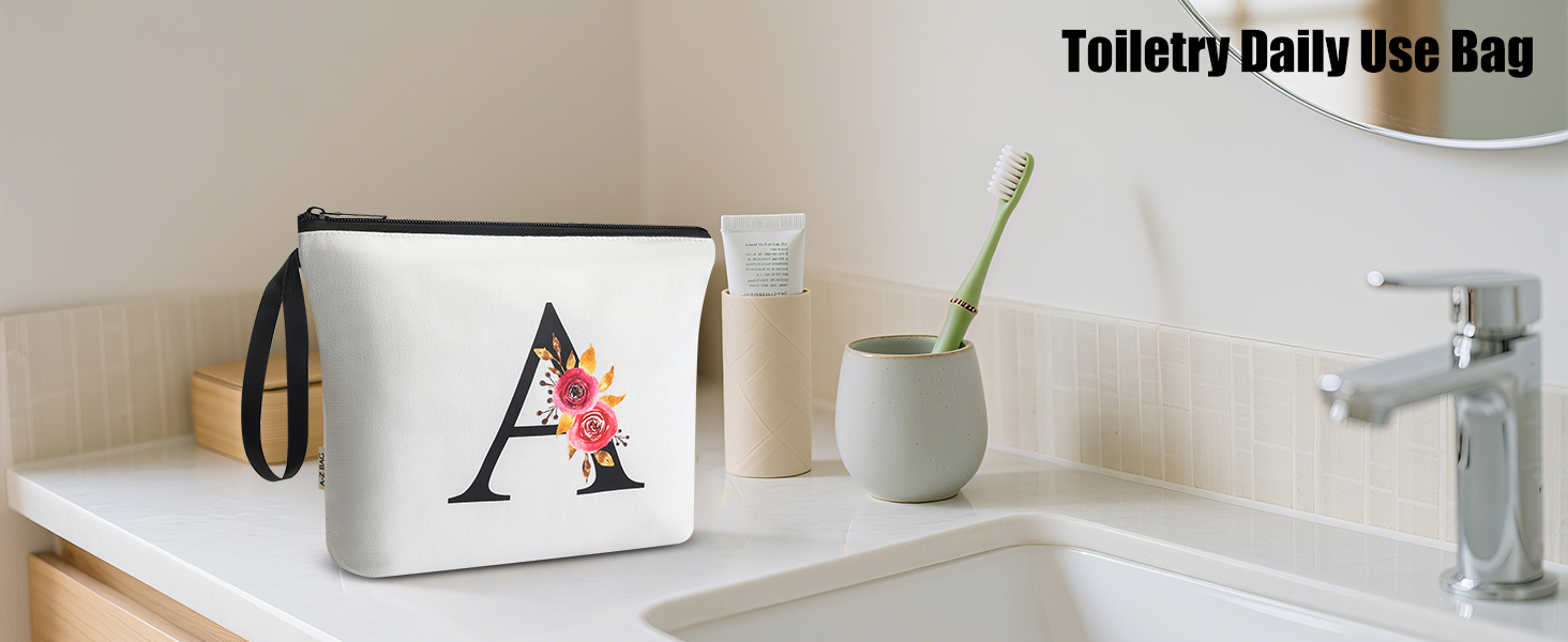 Text reads 'Toiletry Daily Use Bag'. White toiletry bag with decorative letter 'A' in red displayed on bathroom counter next to sink and small plant.