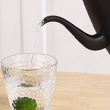 Théière noire versant du liquide dans une tasse en verre texturé sur une surface en bois, vue partielle montrant le bec