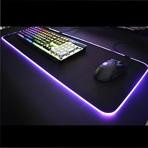 Gaming setup featuring RGB backlit keyboard and mousepad with purple LED lighting effects. Black gaming peripherals on dark surface with dramatic lighting.