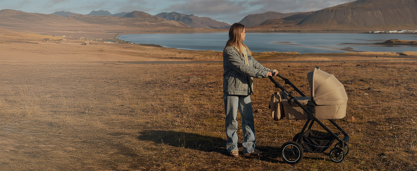 Kinderwagen mit beigem Baldachin auf trockenem Gelände in der Nähe eines Sees. Eine Person in Jeans steht daneben, mit Bergen im Hintergrund