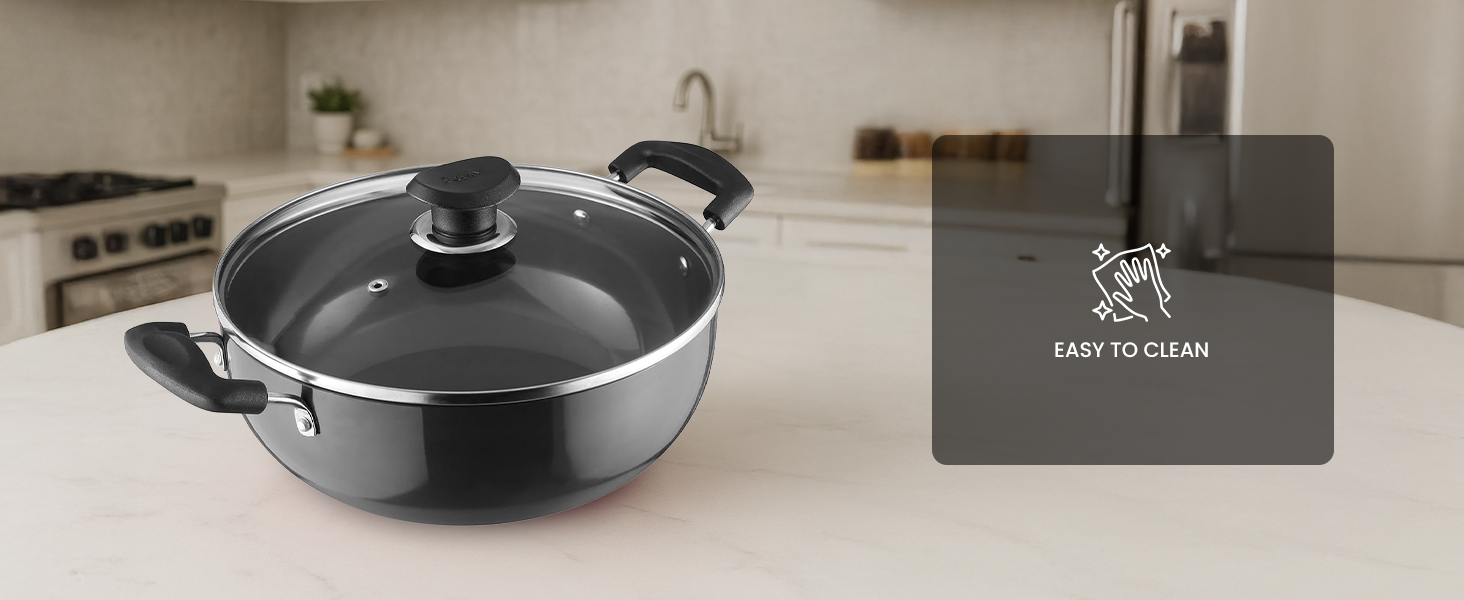 Round black non-stick cooking pan with glass lid and two handles, displayed on white counter next to branded card.