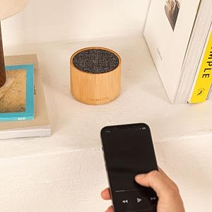 Una mano que sostiene un smartphone negro cerca de un altavoz cilíndrico de madera en un estante blanco con libros y objetos decorativos.