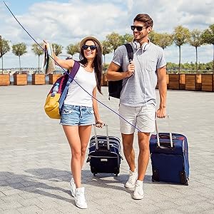 Two people pulling luggage on a sunny outdoor walkway. They're dressed casually, wearing sunglasses, and carrying backpacks alongside their rolling suitcases.