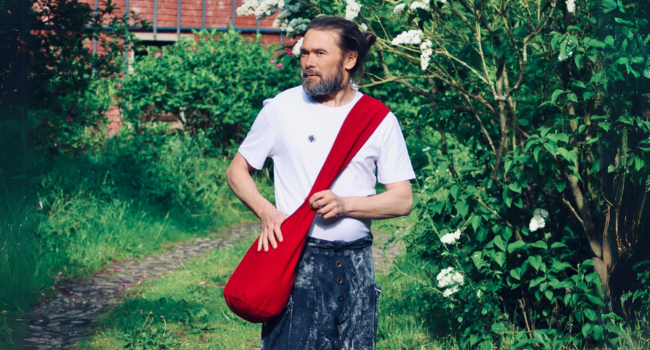 Eine Person, die ein weißes T-Shirt trägt und eine lange rote zylindrische Tasche oder einen Koffer über der Schulter in einer Gartenumgebung trägt.