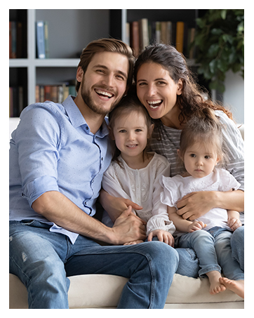 A family seated together on a couch, smiling at the camera. The image includes two adults and two young children.