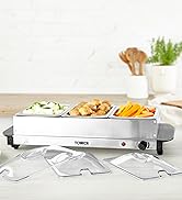 White electric warming tray or buffet server on marble countertop with various food items displayed. Green plant and serving accessories visible in background.