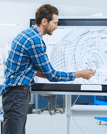 Person in blauem kariertem Hemd, die große technische Diagramme an einem modernen Büroarbeitsplatz überprüft oder darauf zeichnet.