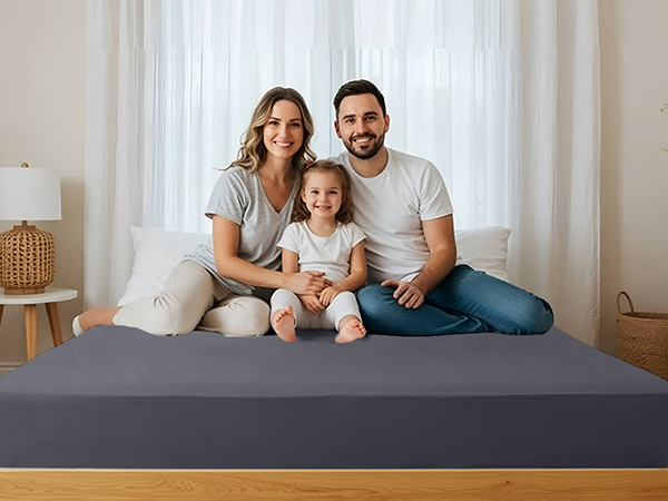 A gray mattress on a wooden bed frame. Three people are seated on the mattress, with white curtains visible in the background.