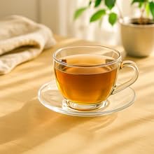 Tasse à thé et soucoupe en verre transparent remplies de thé ambré, photographiées à la lumière naturelle du soleil projetant de douces ombres sur une surface beige.