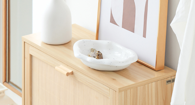 Armario de madera con acabado de color claro, con una superficie superior lisa. En la parte superior hay un jarrón de cerámica blanca y un cuenco poco profundo, junto a una obra de arte enmarcada apoyada contra la pared
