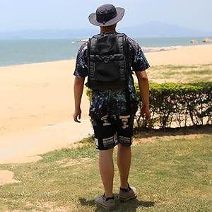 Schwarzer Rucksack, den eine Person am Strand trägt. Breitkrempiger Hut, geblümtes Hemd und Shorts sichtbar. Ozean und Sandstrand im Hintergrund.