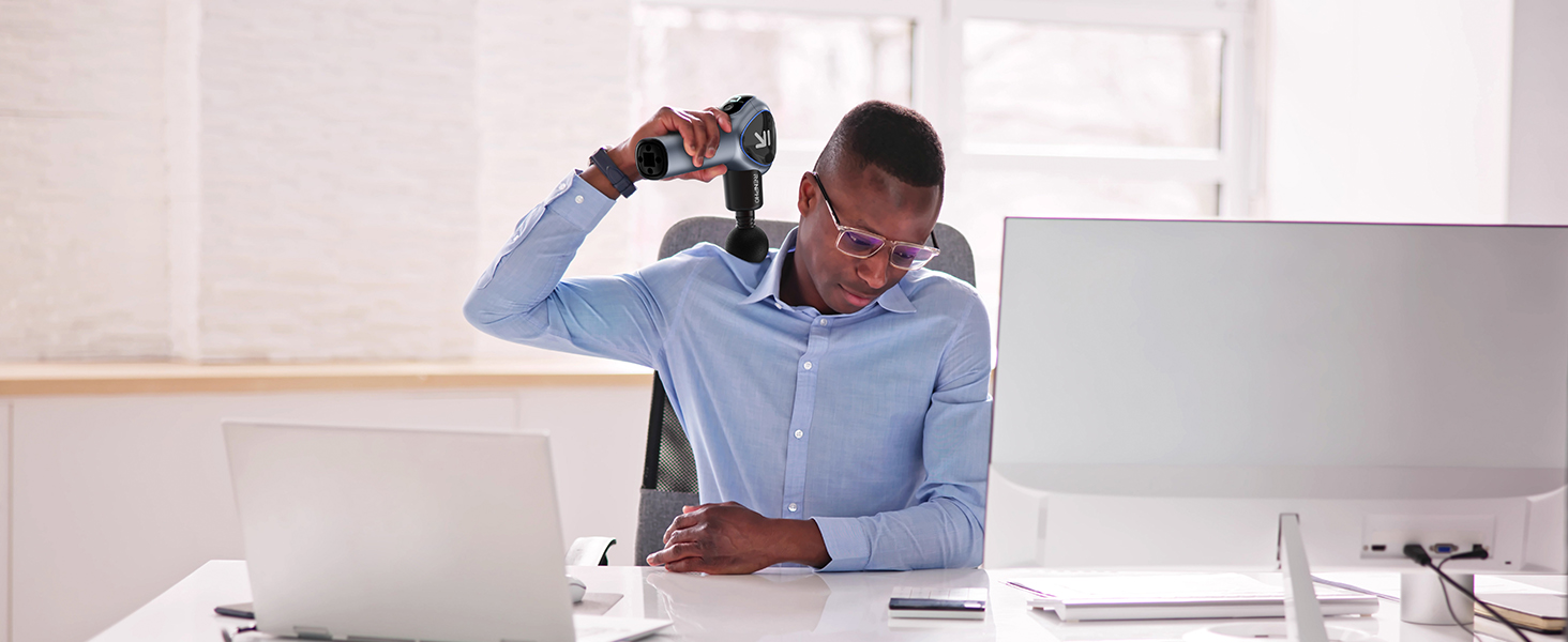 Hombre con camisa azul en el escritorio que usa un taladro eléctrico inalámbrico mientras mira las pantallas de las computadoras, demostrando el uso de herramientas en la oficina.