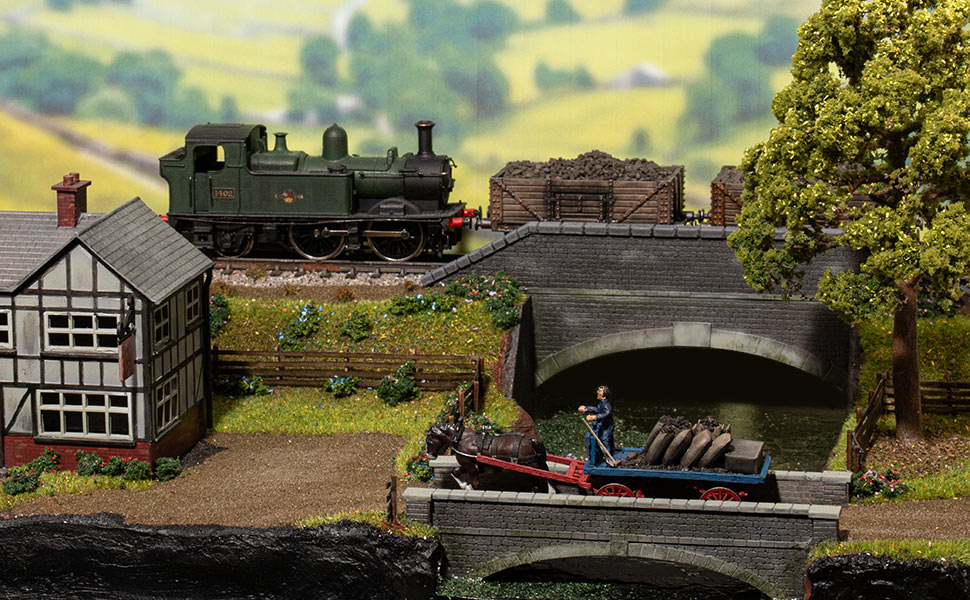 Detailed model railway scene featuring a steam locomotive, Tudor-style building, stone bridge, and canal boat in a rural landscape setting.