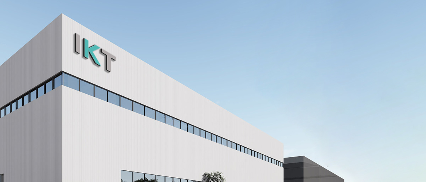 Gran edificio industrial blanco con ventanas teñidas de azul y el logotipo «IKT» en la fachada, sobre un cielo azul claro