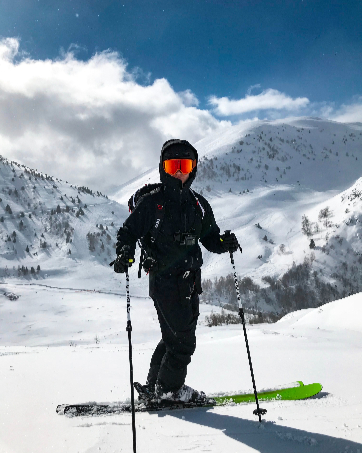 Esquiador con ropa de invierno negra contra un telón de fondo montañoso cubierto de nieve, que usa bastones de esquí mientras esquía cuesta abajo en un día soleado con cielos azules y nubes blancas.