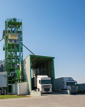 Industrielle Verladeanlage mit grüner Metallstruktur und zwei Sattelschleppern, die unter einer überdachten Ladefläche vor blauem Himmel geparkt sind.