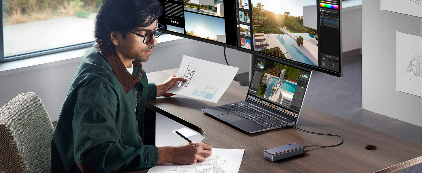 Personne au bureau utilisant plusieurs écrans et un ordinateur portable pour la retouche photo. L'espace de travail comprend une tablette graphique, un clavier et des croquis sur papier dans une pièce ensoleillée avec vue sur la fenêtre