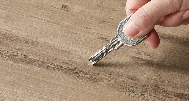 Detailný záber ruky škriabajúcej kľúčom drevený povrch, čo pravdepodobne naznačuje poškodenie alebo opotrebovanie podlahy.