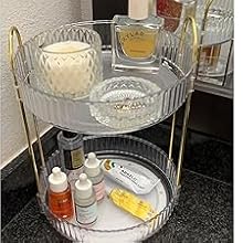 Two-tier gold metal organizer holding various bathroom items including candles, lotions, and a scale on a dark countertop.