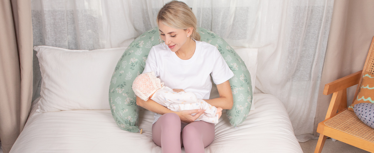 Frau sitzt auf dem Bett mit einem großen, mintgrünen U-förmigen Stillkissen, das um ihre Taille gewickelt ist. Weiße Vorhänge und Holzmöbel sind im Hintergrund sichtbar