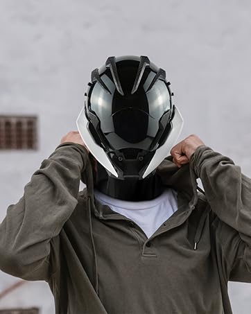 Black motorcycle helmet with glossy finish and angular design. Person wearing olive-green jacket putting on the helmet against a gray background.