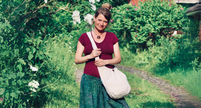 Frau in burgunderfarbenem Top und grünem Rock, die auf einem Naturpfad läuft und eine helle Schultertasche trägt. Umgeben von üppigem Grün