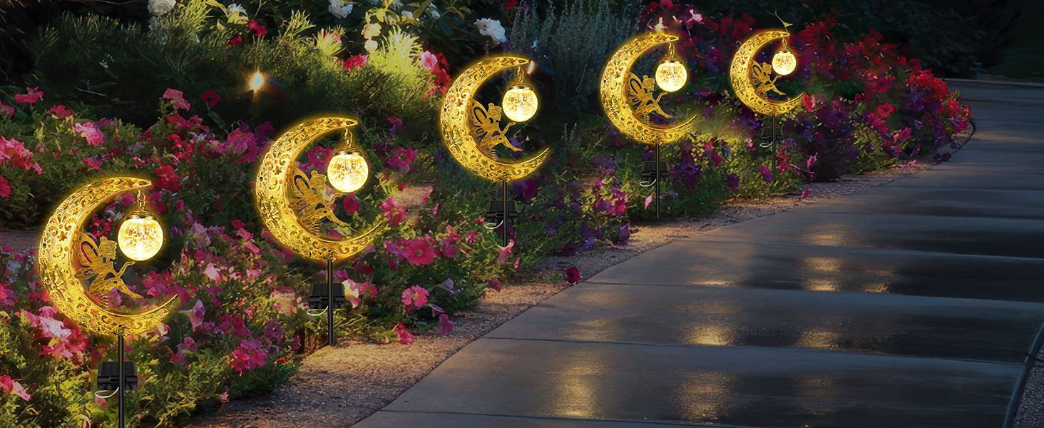 Decorative outdoor solar lights shaped like crescent moons with stars, illuminated in golden color. Placed along a garden path with colorful flowers, creating a magical nighttime ambiance.