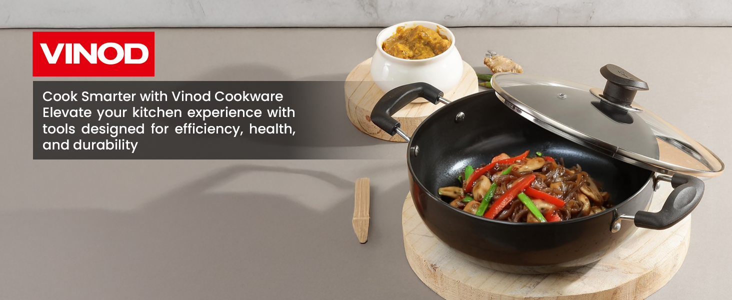 Vinod brand black non-stick cooking pot with glass lid, featuring dual handles, shown cooking stir-fried vegetables.
