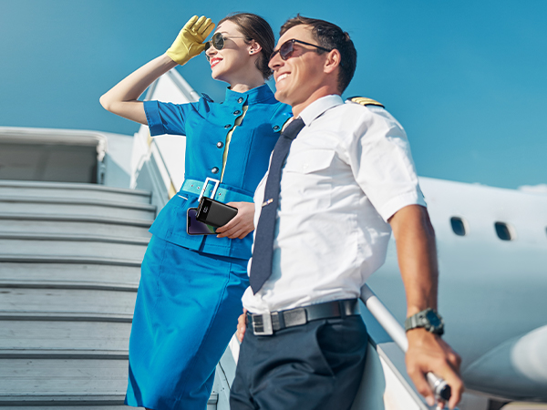 Flight attendant in blue uniform and pilot in white shirt standing outside an aircraft, looking into the distance with hand shielding eyes.