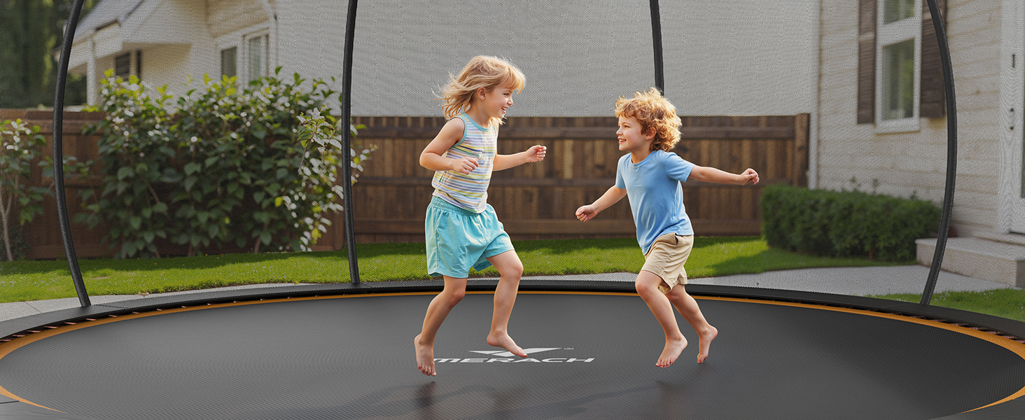 Large round trampoline with black jumping surface. Two children jumping barefoot on the trampoline in a backyard setting with bushes and house visible.