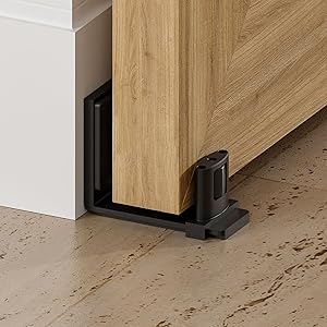 Black sliding door hardware installed at base of wooden door. L-shaped floor guide with roller mechanism visible on tiled floor against white baseboard.