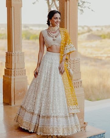 Woman wearing white embroidered lehenga with crop top and layered necklace. Yellow dupatta visible. Standing in ornate archway overlooking landscape.