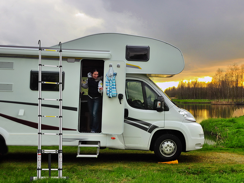 Large white motorhome parked by a lake at sunset. Person visible in doorway. Ladder attached to side, scenic background with trees and water.