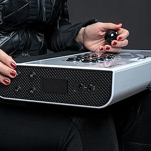 Des mains avec du vernis à ongles rouge actionnent une console de jeu rectangulaire blanche dotée de plusieurs boutons et d'un joystick