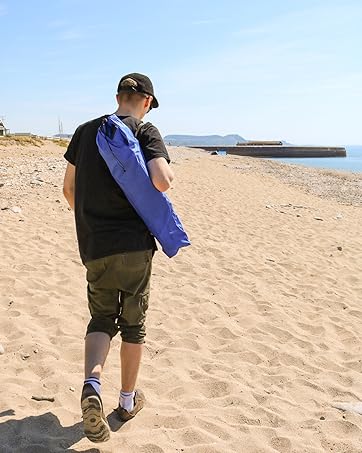 young man walks along the beach, carrying a low beach chair in its bag