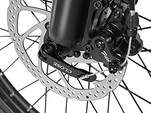 Close-up of a bicycle disc brake system showing a metallic rotor with ventilation holes mounted to the wheel hub, with black brake caliper assembly.
