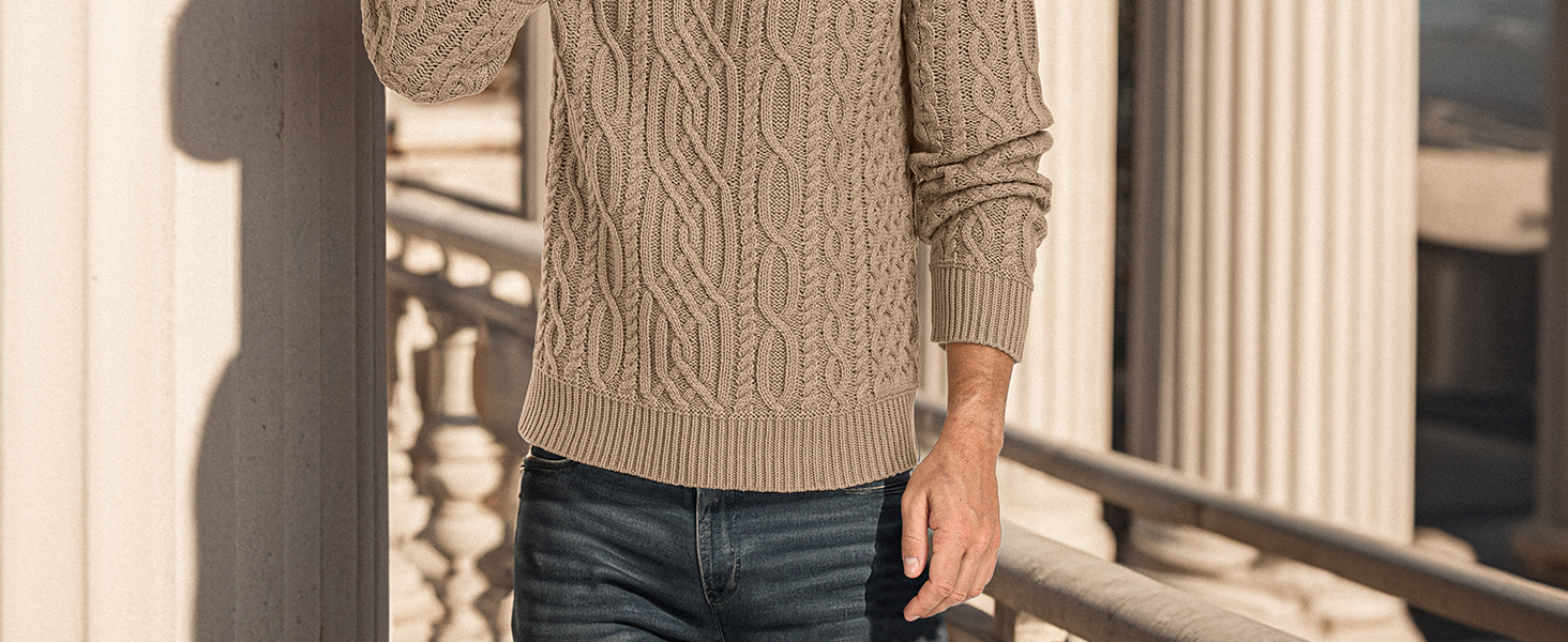 Person wearing beige cable-knit sweater and blue jeans, standing near a stone or concrete pillar. Only torso and arms visible.