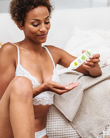 Woman in white undergarments sitting on bed, applying lotion from a white and green bottle to her arm. Demonstrates skincare product usage.