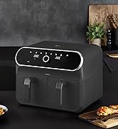Black digital air fryer with LED display panel shown in kitchen setting with prepared food items on plates beside it.